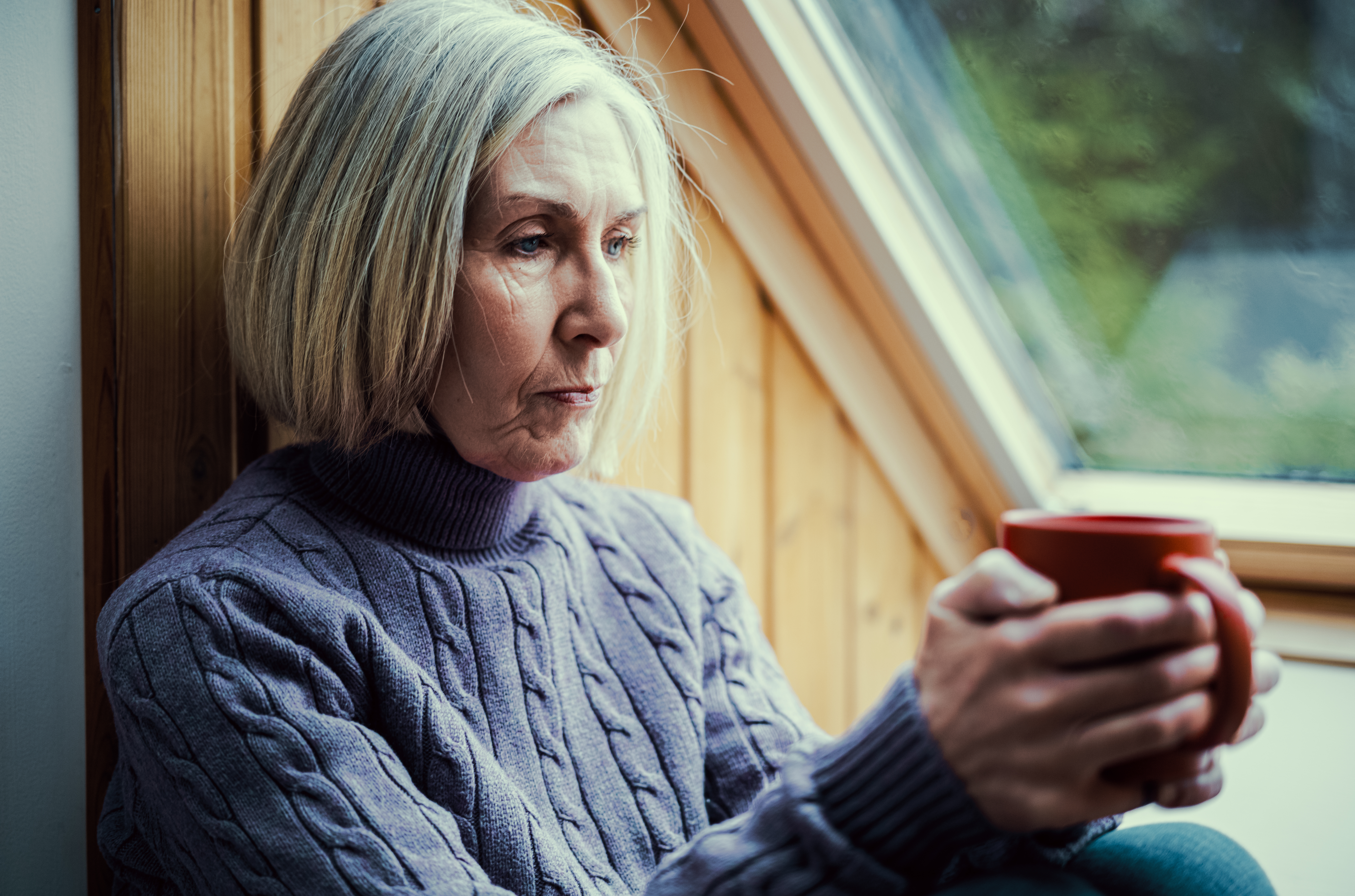 A pensive older woman sits alone by a window holding a red mug. Untreated hearing loss can increase feelings of isolation and is the number one modifiable risk factor for dementia.